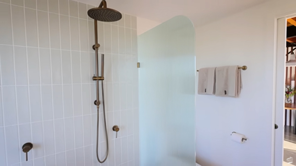 Moderní sprchový kout s mosaznou sprchovou hlavicí a skleněnou zástěnou v minimalistické koupelně.