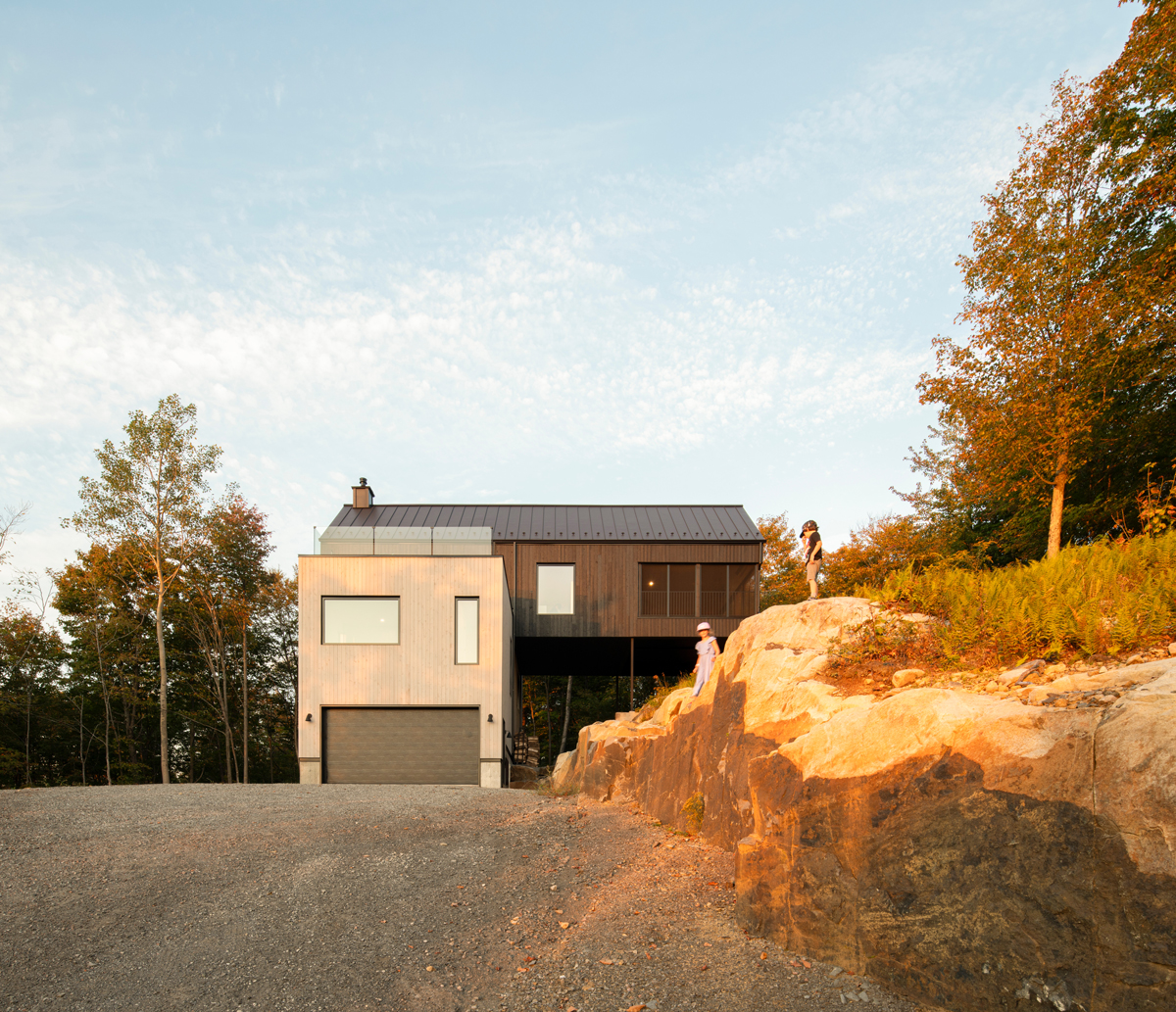 Moderní dům s tmavou střechou zasazený do skalnatého svahu, nasvícený teplým podvečerním světlem.