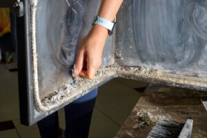 Žena v oranžových rukavicích čistí sklo krbových dvířek popelem a houbičkou.