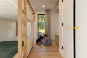 Dětský pokoj se dvěma postelemi a oknem do zahrady, kde si děti hrají na podlaze.