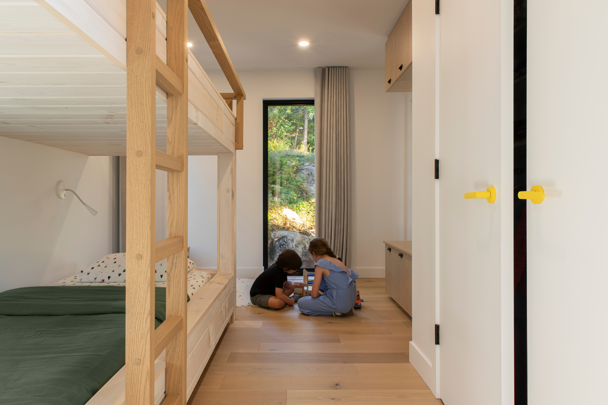 Dětský pokoj se dvěma postelemi a oknem do zahrady, kde si děti hrají na podlaze.