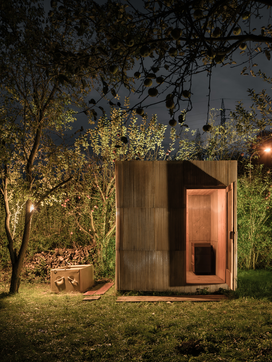 Malá dřevěná sauna z europalet a sklolaminátu zasazená do přírodní zahrady.