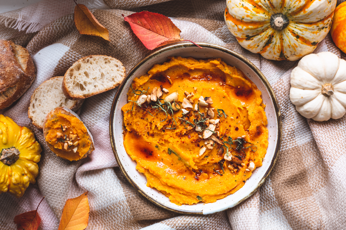 Krémový dýňový hummus s nasekanými ořechy a čerstvými bylinkami, podávaný s křupavým chlebem.