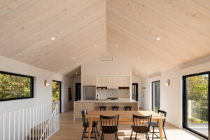 Světlá jídelna s vysokým stropem obloženým dřevem, jednoduchým stolem a černými židlemi.