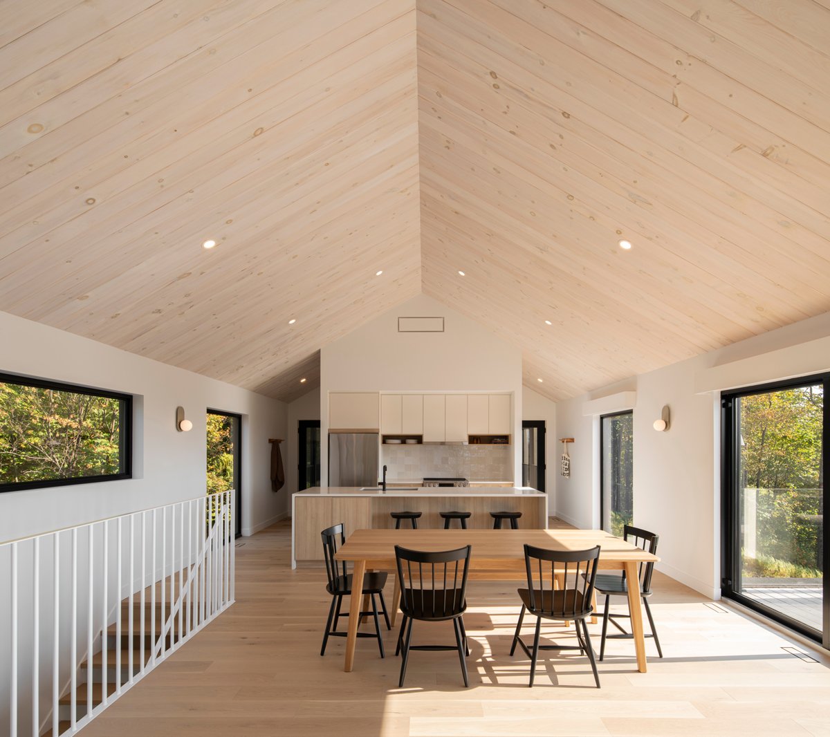 Světlá jídelna s vysokým stropem obloženým dřevem, jednoduchým stolem a černými židlemi.