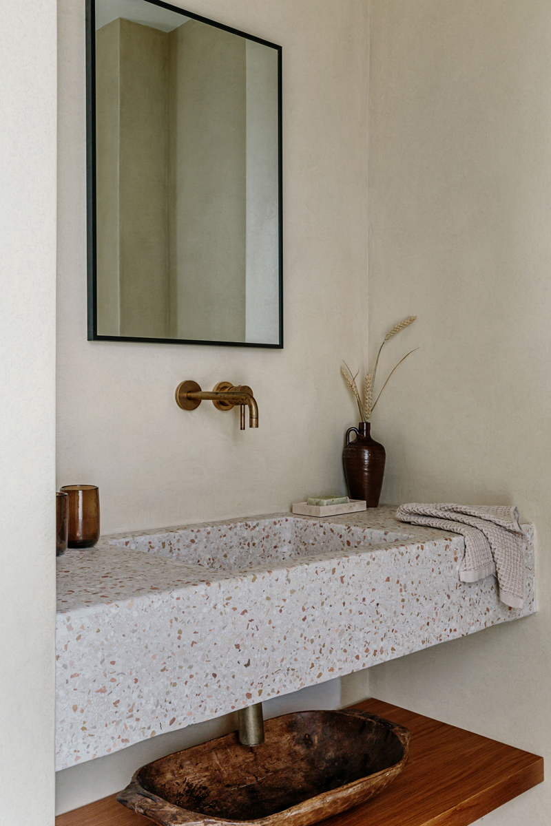 Minimalistická koupelna s terrazzo umyvadlem, bronzovou baterií a rustikálními doplňky.