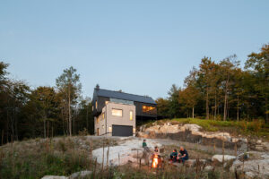 Rodina sedící u ohně před moderním domem zasazeným do svahu, obklopeným lesem.