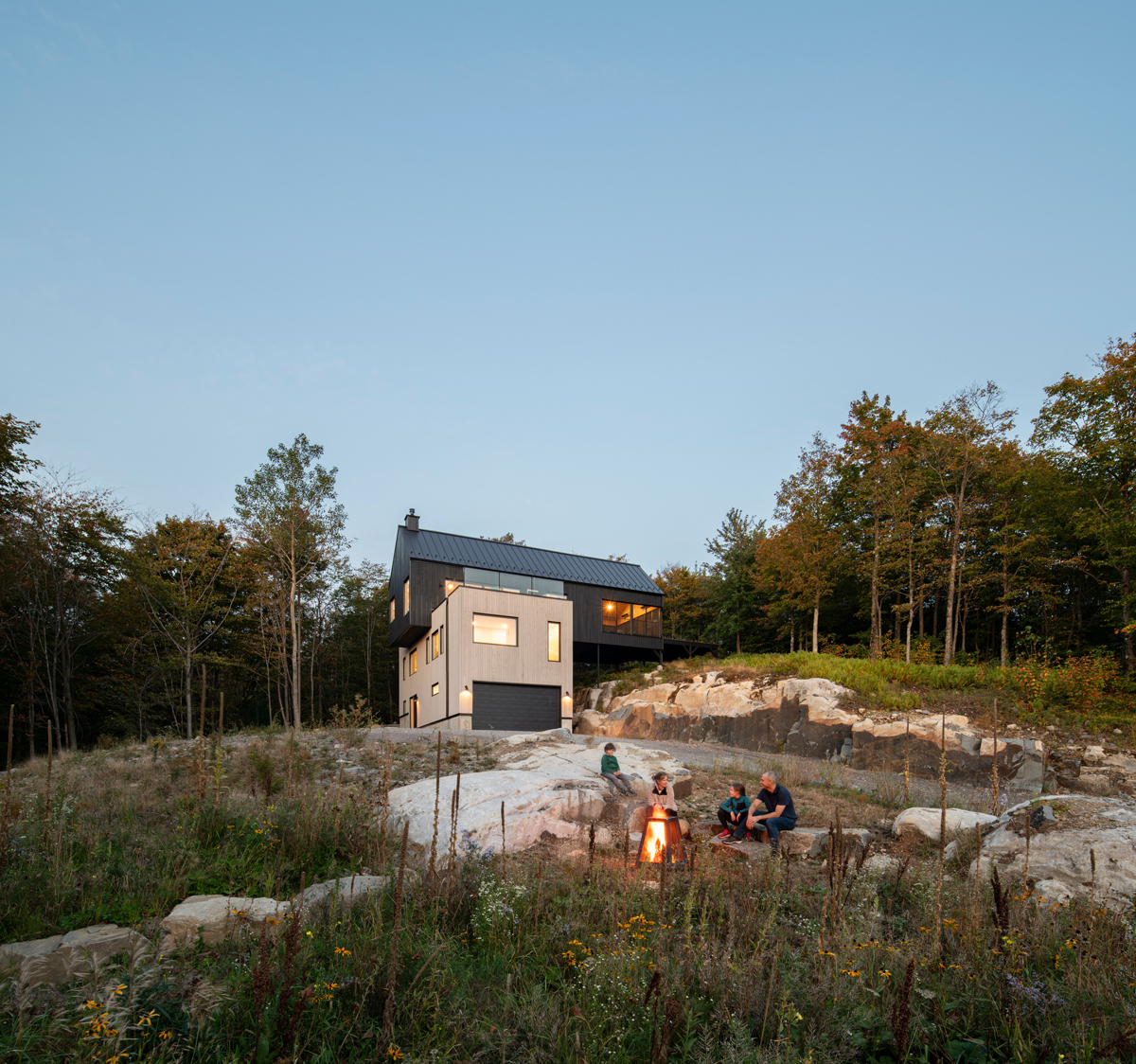 Rodina sedící u ohně před moderním domem zasazeným do svahu, obklopeným lesem.