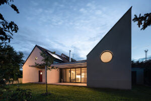 Osvětlený rodinný dům v Křenovicích při soumraku, s prosklenou fasádou a zelenou střechou.