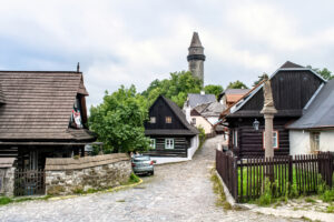 Ulička s dřevěnými roubenkami a kamenným sloupem ve Štramberku s rozhlednou Trúba v pozadí.