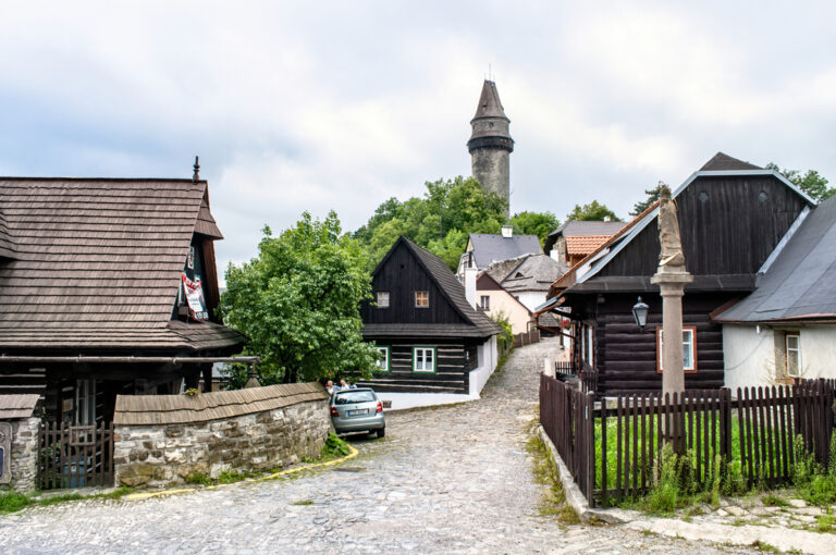 Ulička s dřevěnými roubenkami a kamenným sloupem ve Štramberku s rozhlednou Trúba v pozadí.
