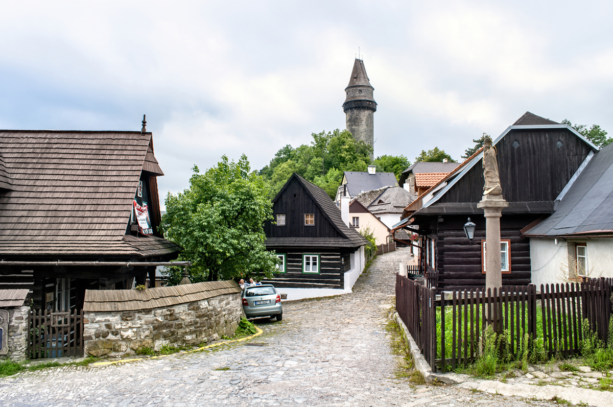 Ulička s dřevěnými roubenkami a kamenným sloupem ve Štramberku s rozhlednou Trúba v pozadí.