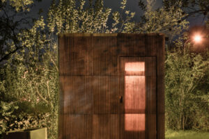 Osvětlená sauna z vlnitého sklolaminátu a dřevěné konstrukce v zahradě během noci.