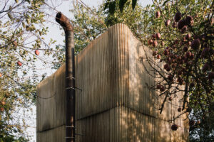 Kovový komín venkovní sauny s fasádou z vlnitého sklolaminátu a dřevěného roštu, zasazené mezi ovocné stromy.
