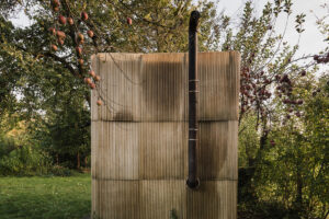 Venkovní sauna z recyklovaných materiálů v parku, obklopená stromy a zahradním osvětlením.