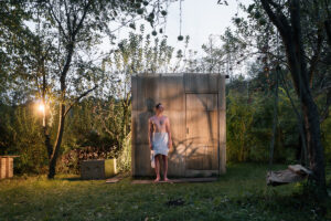 Osvětlená sauna s plechovou fasádou a komínem v městské zahradě při soumraku.