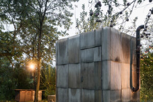 Malá venkovní sauna z recyklovaných materiálů, obložená vlnitým sklolaminátem, zasazená do zahrady.