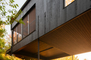 Detail černé dřevěné fasády domu podepřeného subtilními ocelovými sloupy nad kamenitým terénem.