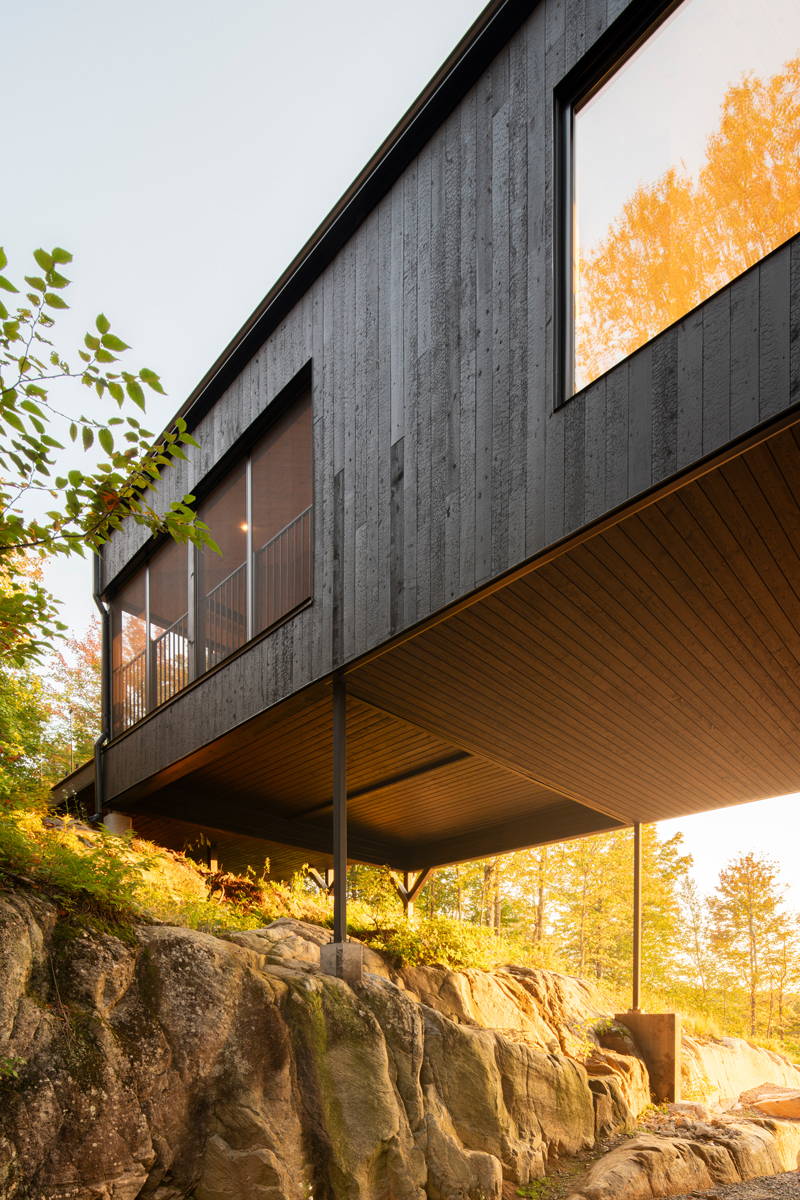 Detail černé dřevěné fasády domu podepřeného subtilními ocelovými sloupy nad kamenitým terénem.