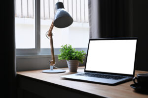 Pracovní koutek s notebookem a stolní lampou u okna, doplněný zelenou rostlinou pro svěží atmosféru.