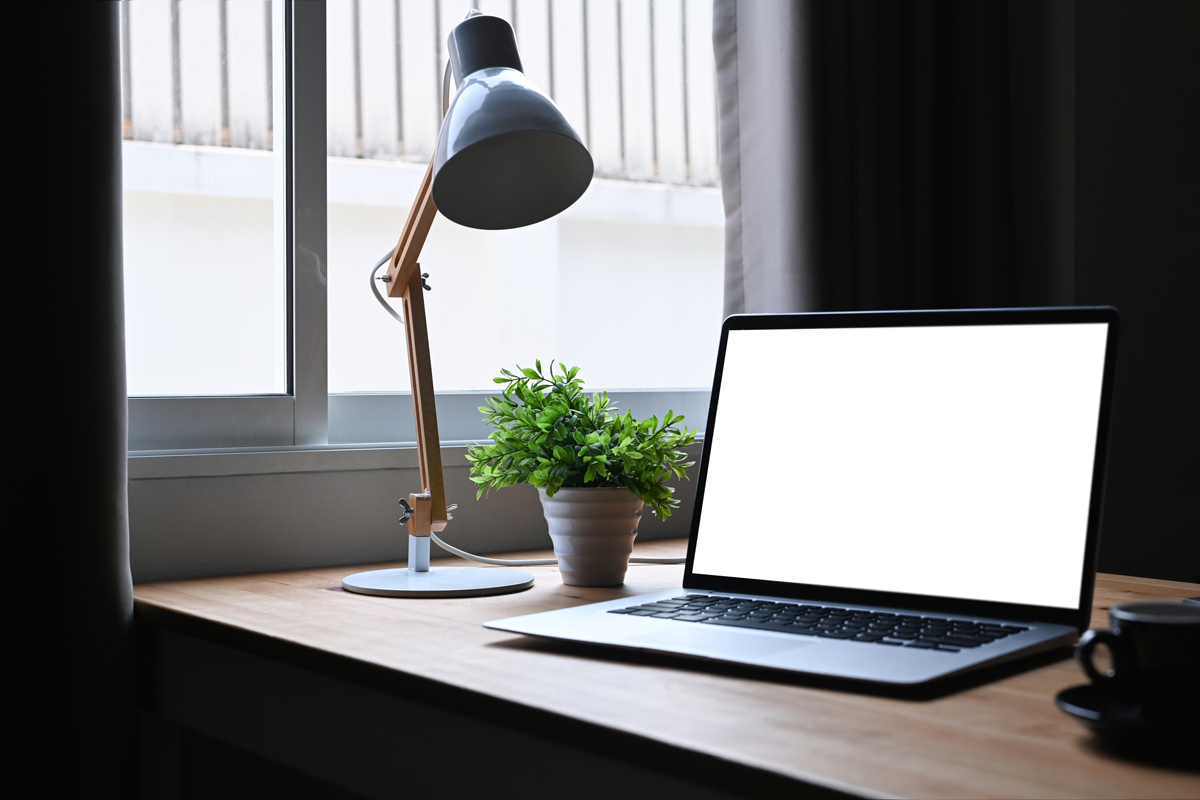 Pracovní koutek s notebookem a stolní lampou u okna, doplněný zelenou rostlinou pro svěží atmosféru.
