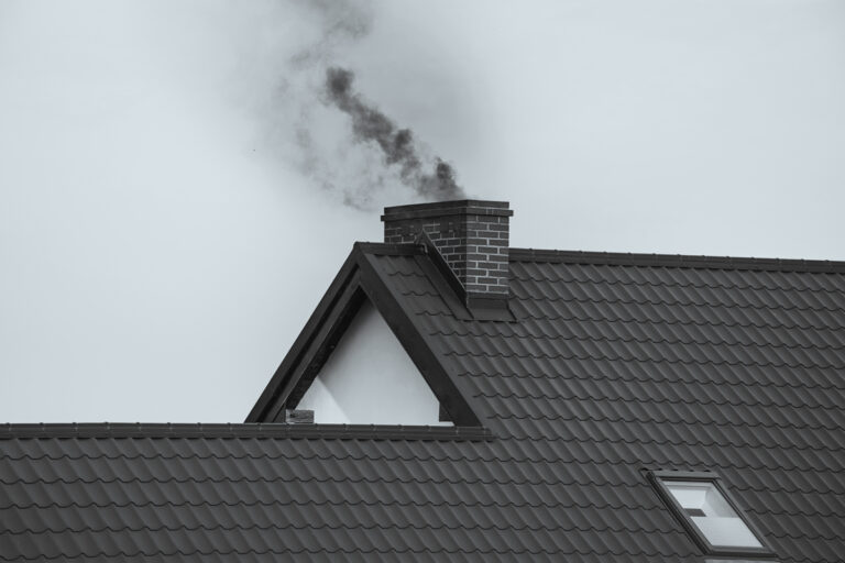 Detail komína s tmavým dýmem stoupajícím nad střechou domu, ukázka neekologického topení tuhými palivy.