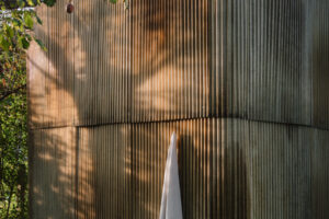 Detail venkovní sauny postavené z vlnitého sklolaminátu, vzniklé z recyklovaných materiálů, s bílým ručníkem zavěšeným na háčku.