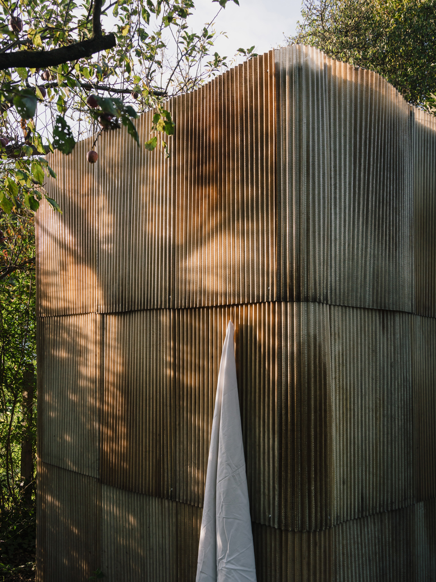 Detail venkovní sauny postavené z vlnitého sklolaminátu, vzniklé z recyklovaných materiálů, s bílým ručníkem zavěšeným na háčku.