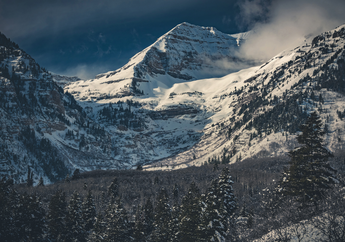 Zasněžené hory Mount Timpanogos s hustými jehličnany a dramatickou oblohou.