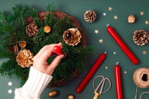 Ruka zdobící přírodní adventní věnec se zelenými větvičkami, červenými svíčkami a šiškami zachycuje proces výroby tradiční vánoční dekorace.