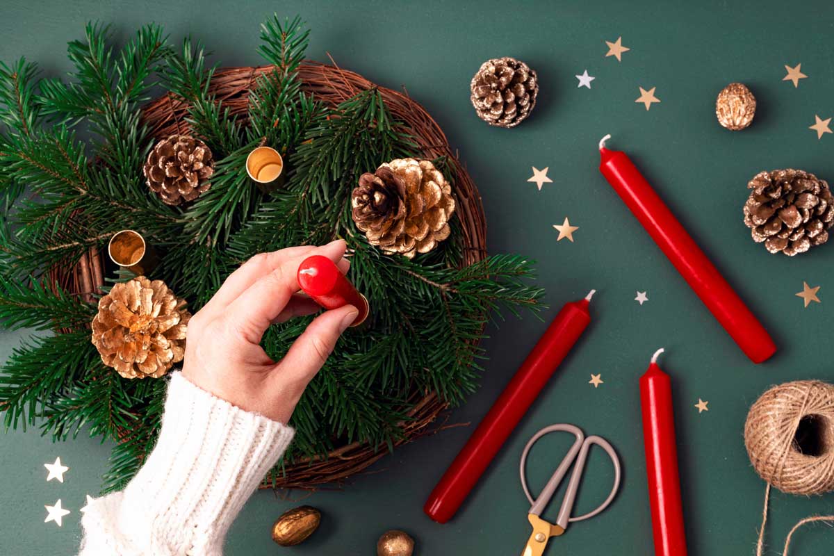 Ruka zdobící přírodní adventní věnec se zelenými větvičkami, červenými svíčkami a šiškami zachycuje proces výroby tradiční vánoční dekorace.