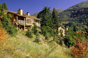 Horská chata obklopená stromy na svahu s výhledem na hory a podzimní zeleň.