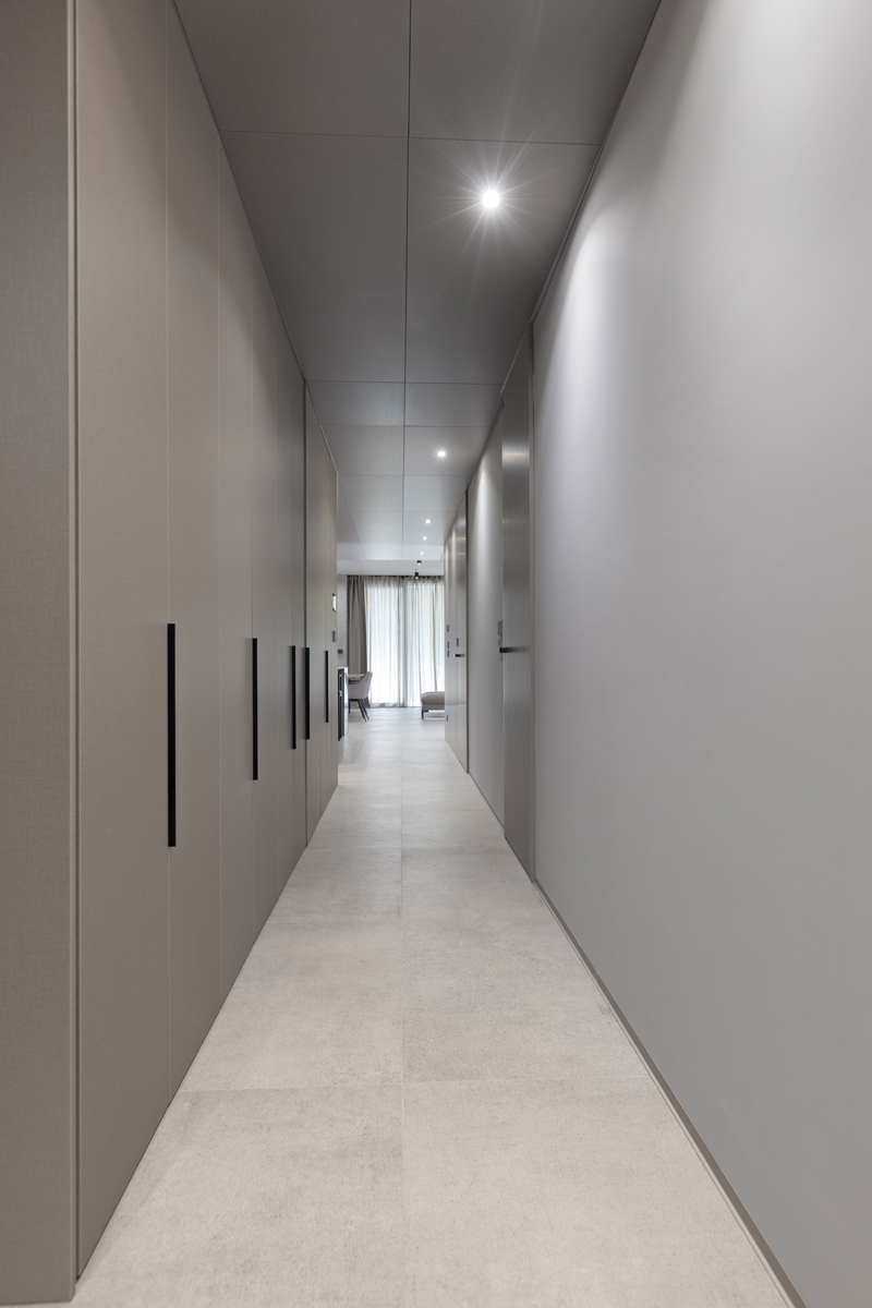 Úzká chodba se zapuštěnými dveřmi a minimalistickým osvětlením v jednotné paletě šedých tónů.