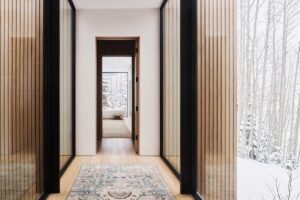 Chodba s prosklenými stěnami a dřevěným svislým obkladem vedoucí do pokoje s výhledem na les.