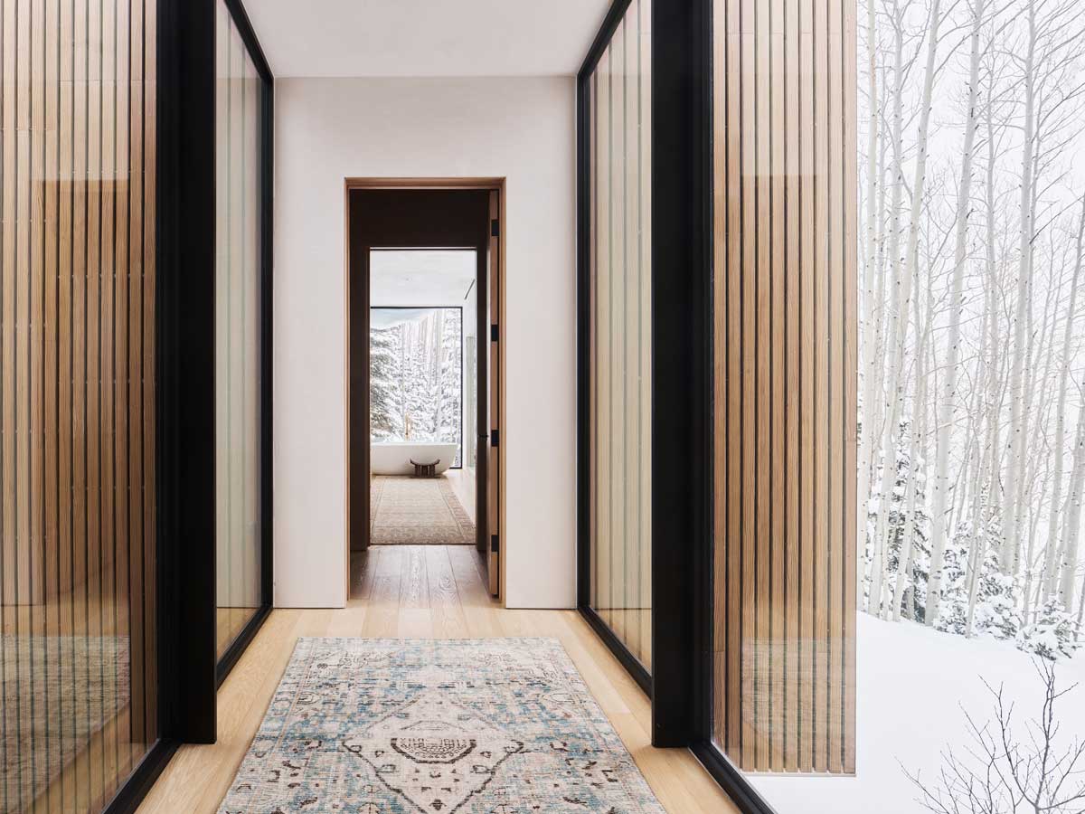 Chodba s prosklenými stěnami a dřevěným svislým obkladem vedoucí do pokoje s výhledem na les.