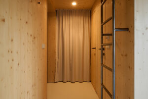 Vertikální žebřík z oceli vede na horní patro s místem na spaní. Kombinace surového kovu a dřeva dodává prostoru industriální akcent.