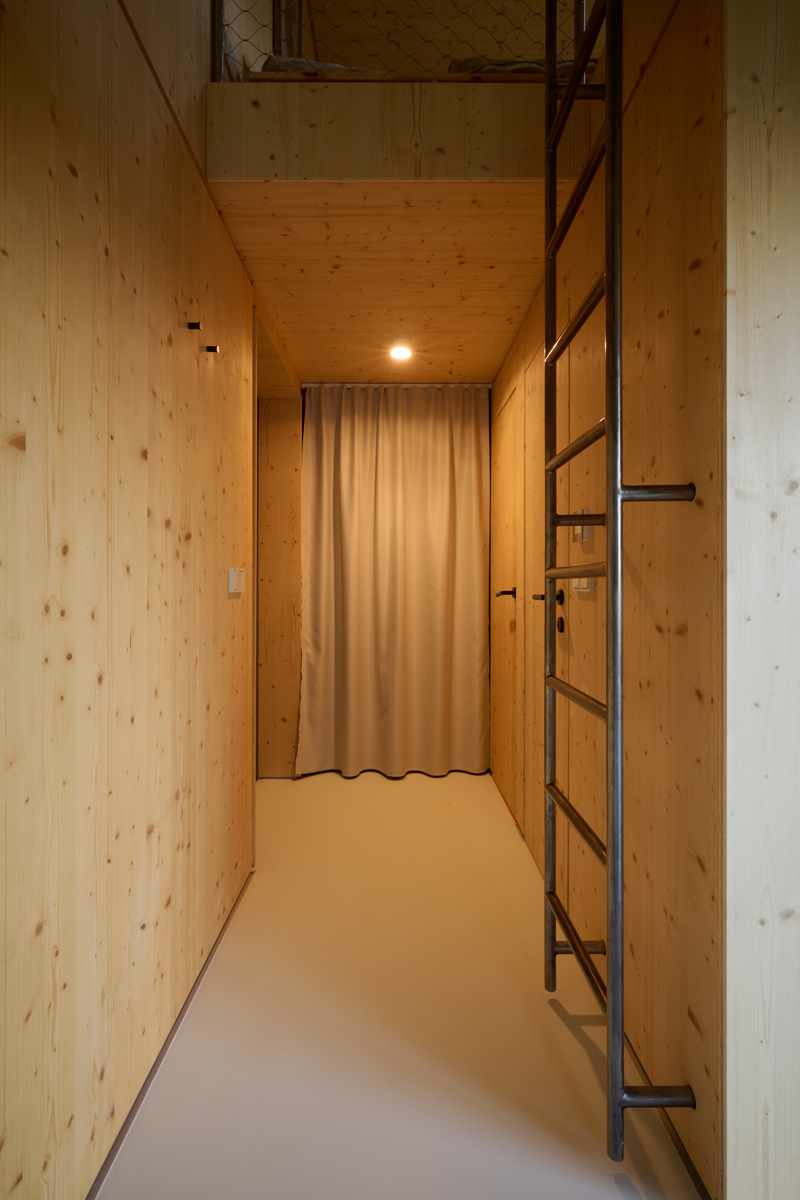 Vertikální žebřík z oceli vede na horní patro s místem na spaní. Kombinace surového kovu a dřeva dodává prostoru industriální akcent.