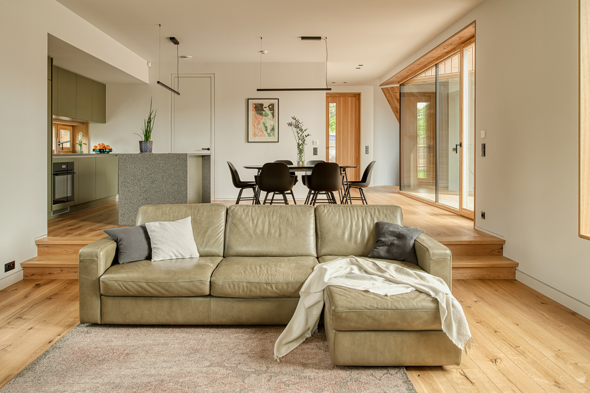 Obývací pokoj s koženou pohovkou, jídelním stolem a výhledem přes velká okna na dřevěnou terasu.