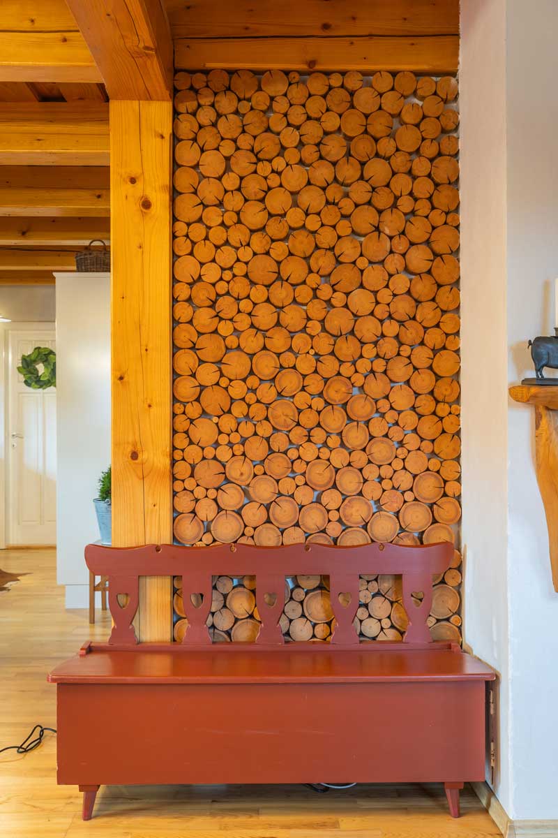 Vnitřní dřevěná stěna s nařezanými špalky jako dekorací, doplněná červenou venkovskou lavicí.