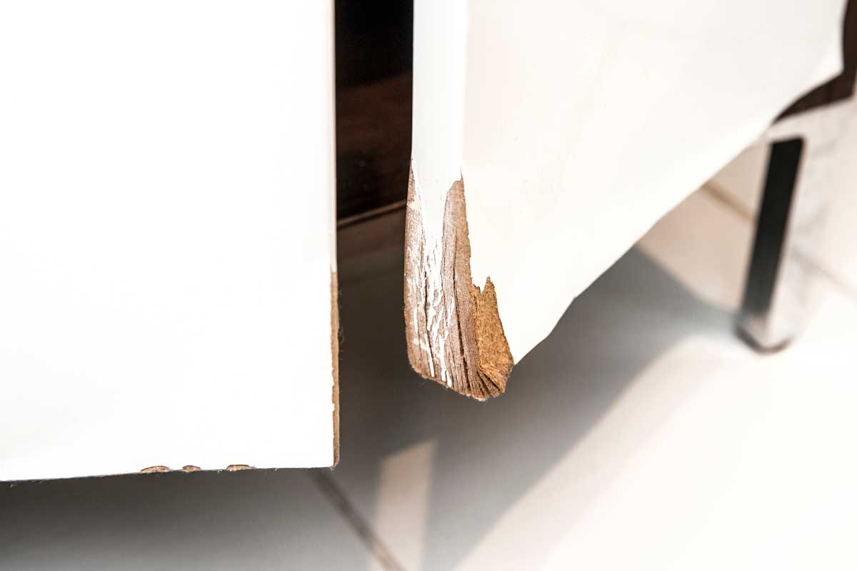 Detail odlupujícího se rohu bílých nábytkových dvířek.