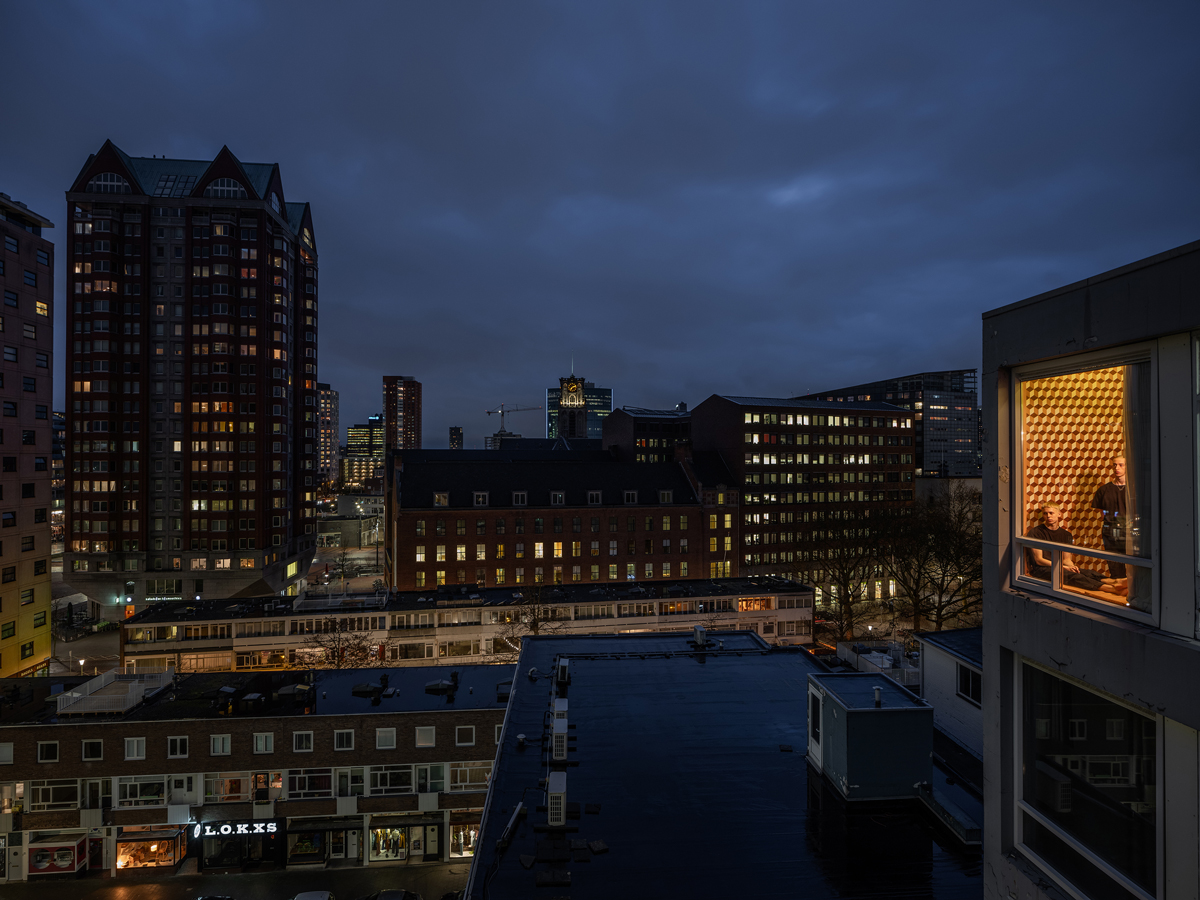 Noční město s osvětleným oknem malého bytu a postavami uvnitř.