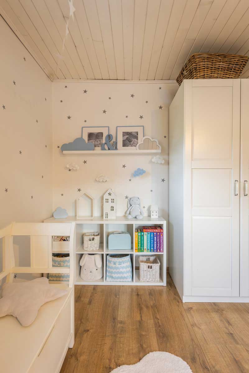 Dětský hrací koutek v jemných tónech s knihami, košíky a dekoracemi v podobě domečků a obláčků.