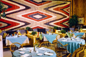 Horská restaurace s dřevěným interiérem, modrými ubrusy a výraznou tkaninou s geometrickým vzorem na stěně.