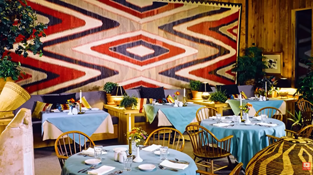 Horská restaurace s dřevěným interiérem, modrými ubrusy a výraznou tkaninou s geometrickým vzorem na stěně.