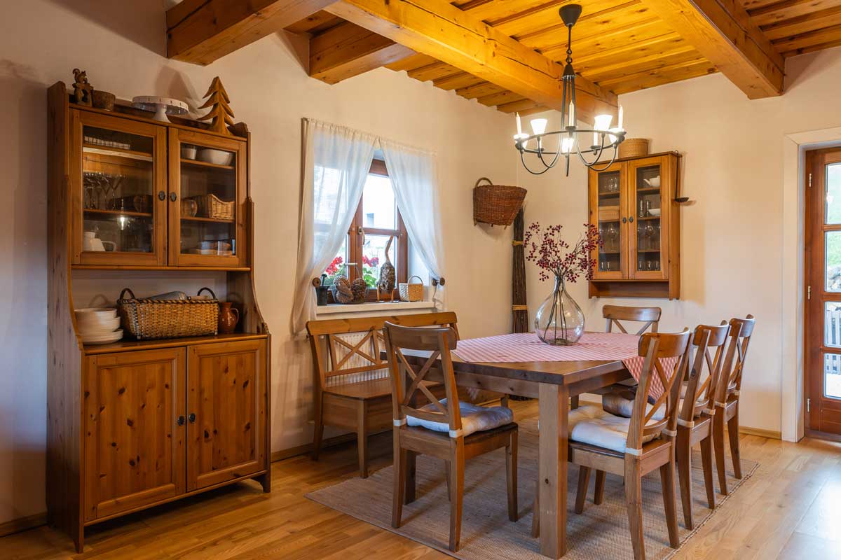 Velký dřevěný jídelní stůl s textilním středem, obklopený židlemi, opticky zateplený dřevěnými trámy na stropě.