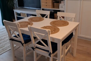 Bílý jídelní stůl se čtyřmi židlemi a kruhovými prostírkami z přírodního materiálu.