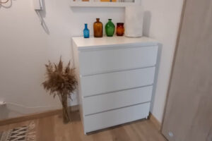 Bílá komoda s barevnými vázami a sušenou travinou v rohu místnosti.