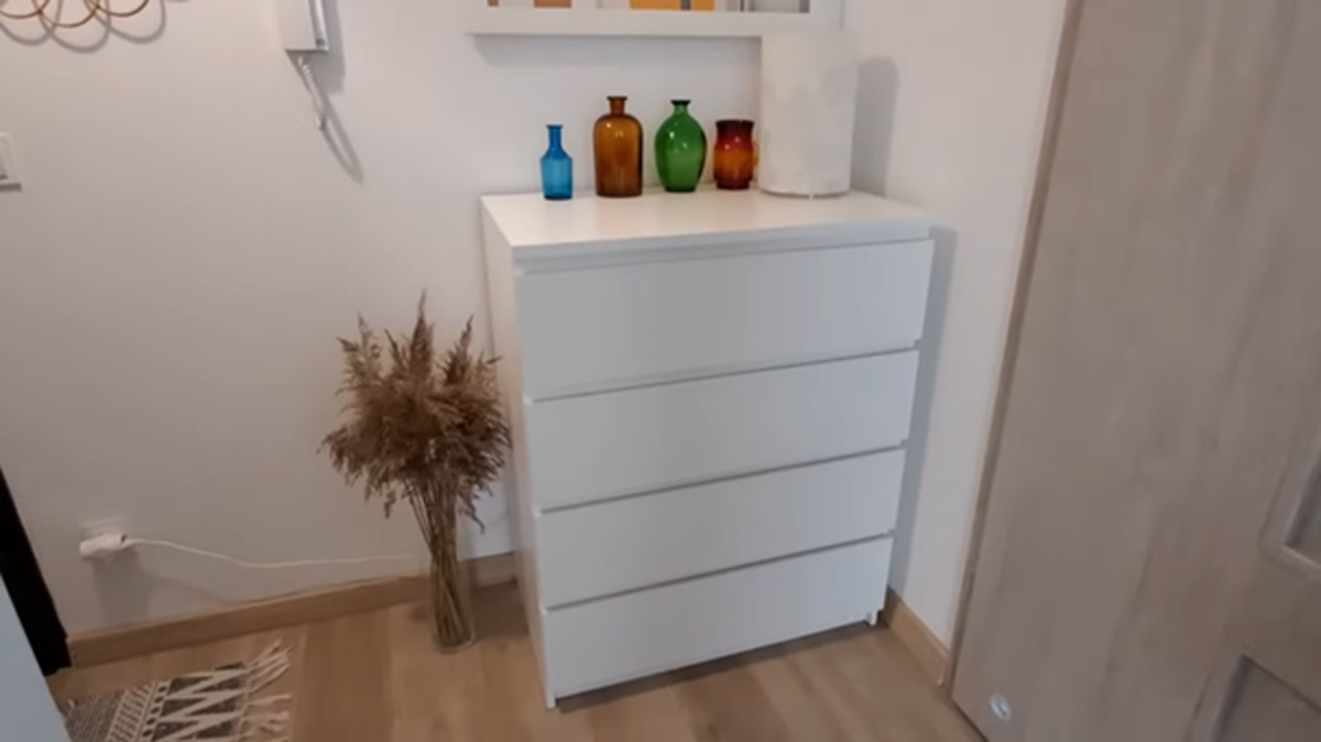 Bílá komoda s barevnými vázami a sušenou travinou v rohu místnosti.