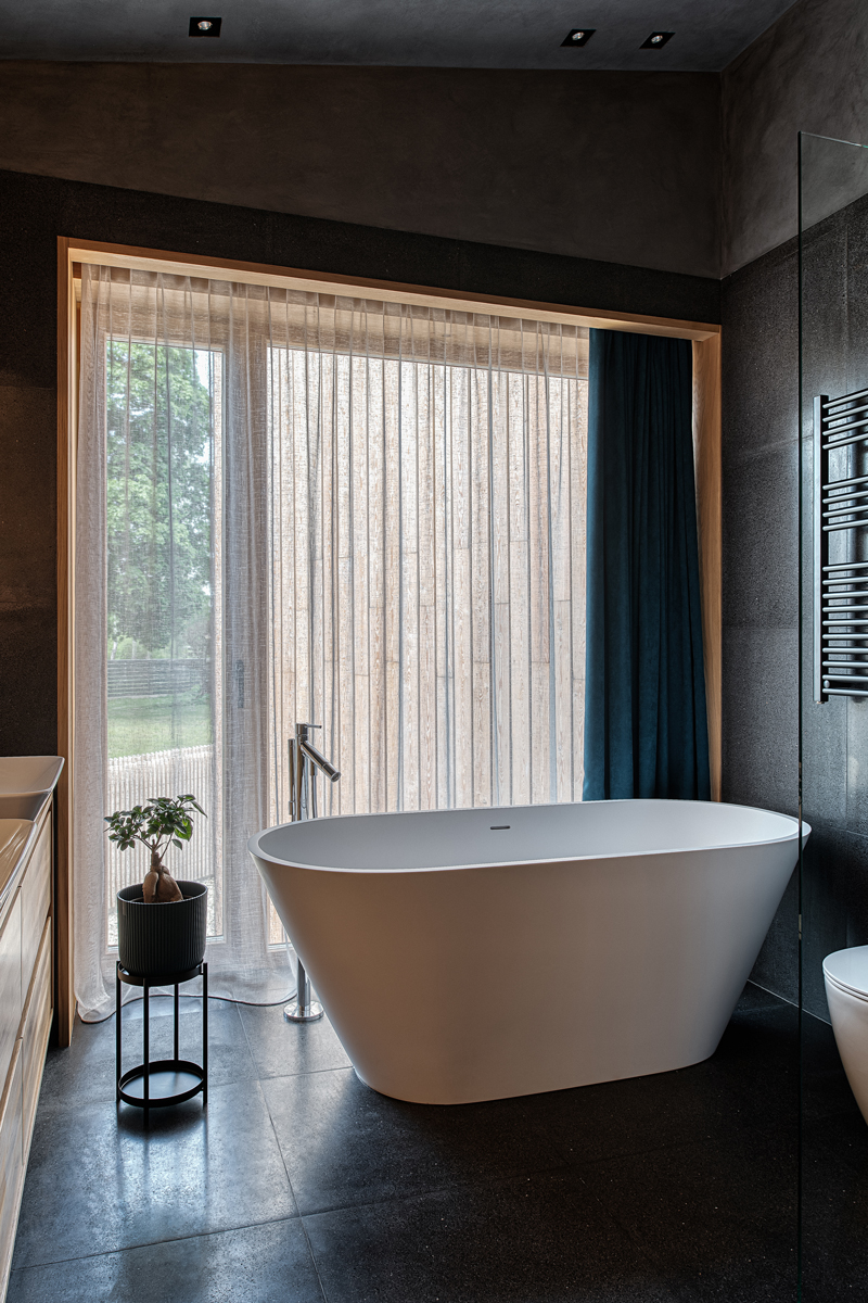 Koupelna s volně stojící vanou, průsvitným závěsem a tmavou podlahou.