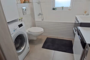 Moderní koupelna s vanou, pračkou a bílým nábytkem v malém bytě.